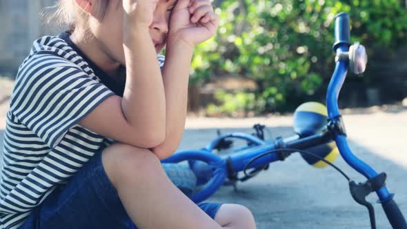 Sad little girl sitting on the ground after falling off her bike at summer park.