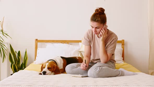 Young Woman Sits on the Bed and Petting Her Dog Beagle While Talking By Phone