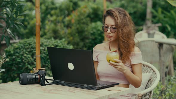 A Young Woman Photographer Drinks a Cocktail While Working with Her Laptop in an Outdoor Cafe