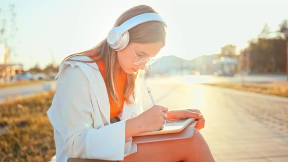 Teenage Girl with Headphones Sitting on Park Bench and Drawing with Stylus on Graphics Tablet