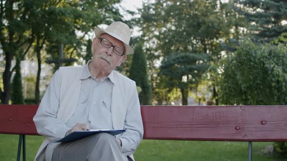 Senior Man Sits Park Bench Thinks Scratches Mustache Nose and Writes on Paper