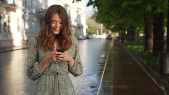 Portrait of Satisfied Young Woman Wearing Dress Smiling and Holding Mobile Phone While Walking