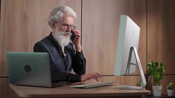 Adult Grayhaired Man Works in the Office at the Computer Typing and Talking on a Mobile Phone an