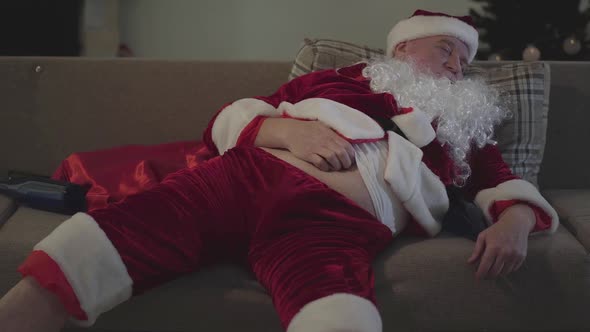 Bad Santa Claus Sleeping on Sofa and Scratching His Belly. Empty Bottle Lying Next To Him on the