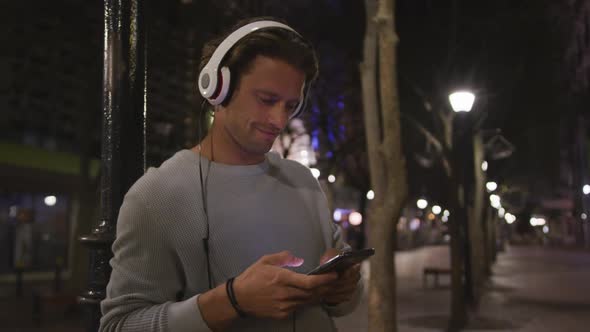 Caucasian male wearing headphones, listening to music, using his phone in the evening