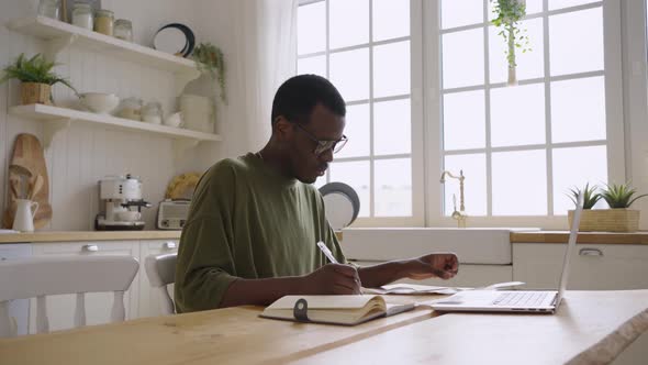 Young Black Man Writes Expenses From Paper Checks in a Diary at Home at a Laptop and Gets Upset at