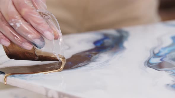 Young Woman Pouring Down the Golden Glittery Epoxy on the Painting From the Plastic Cup