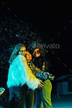 Three happy female friends embracing each other while standing under confetti at night
