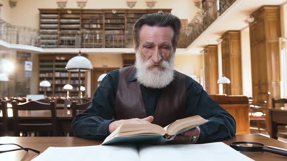 Old Bearded Man which Flipping Through Book Pages Sitting at the Library Table