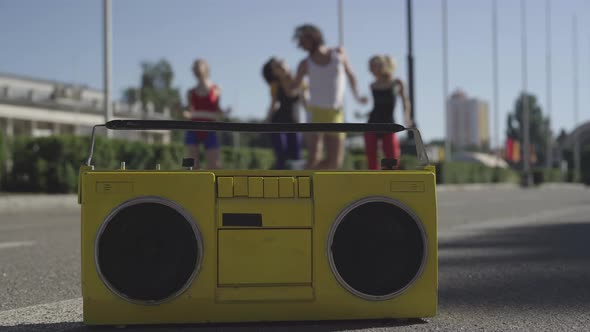 Retro Tape Recorder Standing on Asphalt with Four Caucasian Sportive People Doing Fitness Exercises