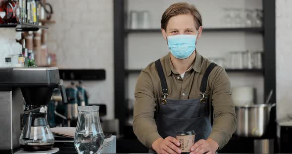 Portrait of Caucasian Handsome Male Barista in Medical Mask and Gloves Giving Cup of Coffee to