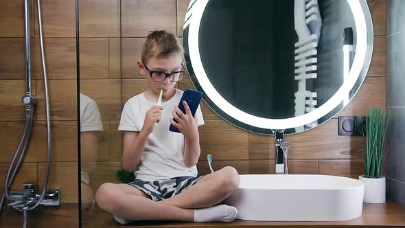 Boy Sitting Near the Bathroom Washbasin and Mirror and Cleaning His Teeth, Browsing Phone Apps