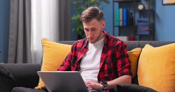 A Handsome College Student Boy is Studying Remotely While Sitting on Couch in Room at alt