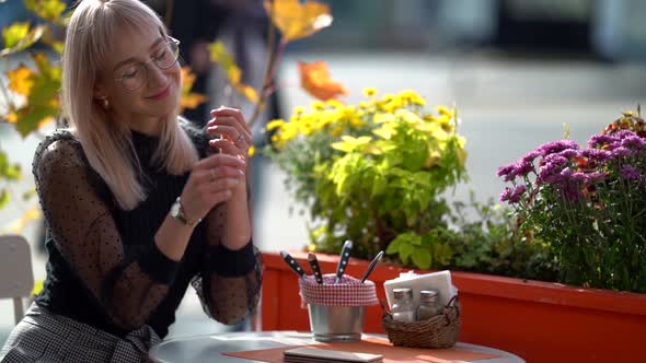 Portrait of a Young Positive Blonde Woman with Glasses Sitting Outside in a Cafe at a Table on a