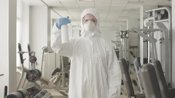 Middle Shot of Serious Woman in Protective Suit and Respirator Spraying Disinfectant in Gym