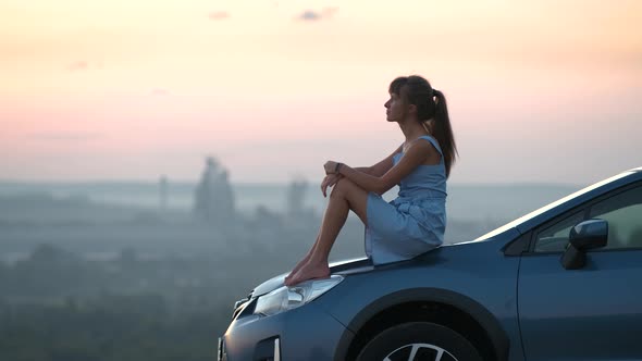 Happy young woman driver in blue dress laying on her car hood enjoying warm summer day.