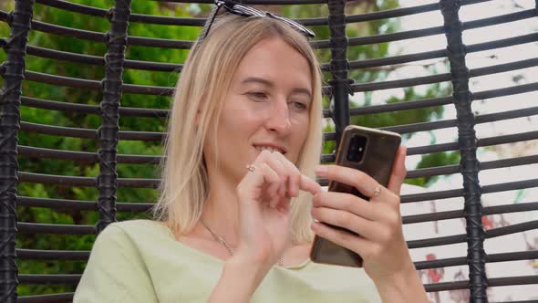 a blonde woman looks at her smartphone and swings in hanging rattan chair