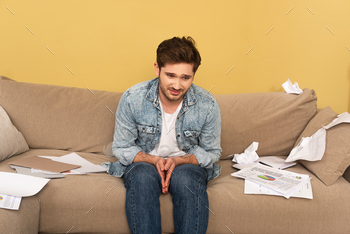 Sad man sitting on couch near crumpled paper and documents with charts