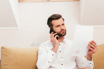 Pensive teleworker talking on smartphone and holding paper on couch