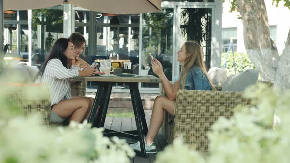 Side View of Young Ladies Friends Talking and Using Smartphones Sitting at Table in Outdoor Cafe