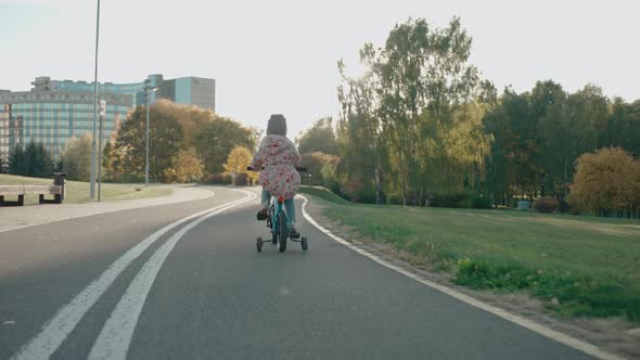 Little Girl Rides a Tricycle Bike in the City Park at Sunset in Sunny Autumn