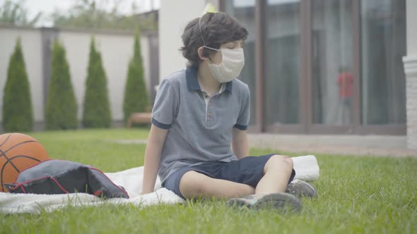 Portrait of Sad Caucasian Boy in Face Mask and Party Hat Sitting on Blanket Outdoors Alone. Upset