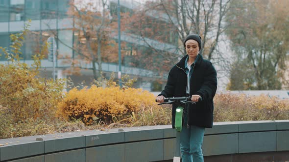 Young Woman in Winter Black Jacket with Hat Riding Electic Scooter in the City Park. Ecological