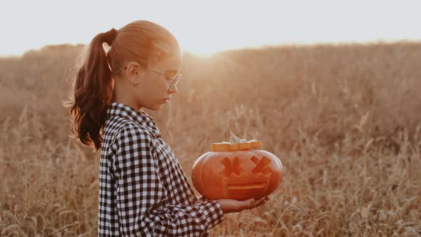 A Teenage Girl Holds a Halloween Pumpkin in Her Hands Against the Backdrop of a Sunset in a Field