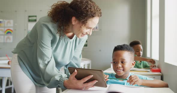 Video of happy caucasian female teacher explaining lesson on tablet to african american boy