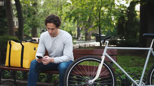 Portrait of Caucasian Delivery Man Sitting on Bench in Central Park and Using Smartphone with Yellow