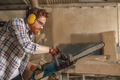 Professional carpenter in glasses is cutting wood with chainsaw on carpentry workshop - PhotoDune Item for Sale