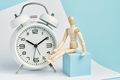 Wooden man sits near a white alarm clock. Concept of watching time, long wait. - PhotoDune Item for Sale