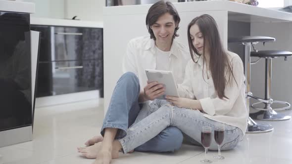 Happy Relaxed Young Couple Sitting on Kitchen Floor with Wine Glasses and Using Tablet. Portrait