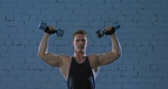 Muscular Young Man Lifts Dumbbells Up Shakes the Muscles of His Arms and Back