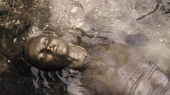 Woman with Skin Painted Golden Dye Is Floating in Shallow with Shiny Gold Pigment