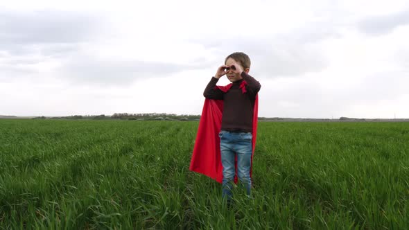 A Small Boy Makes Glasses with His Hands, and Looks Into the Distance. A Child in a Red Superhero