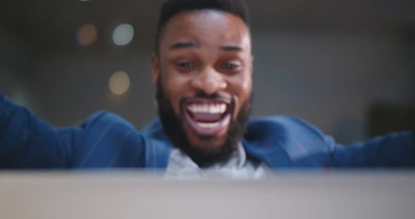 Cheerful Businessman Celebrating with Arms Raised Sitting at Desk in Office and Working on Laptop