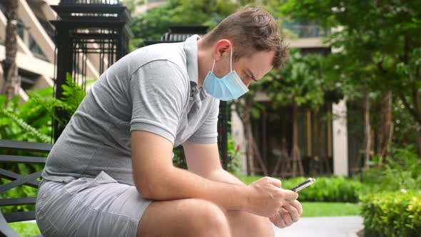 Sad Man Wear Face Mask Sit Hunched Over in Public Park Work with Smartphone