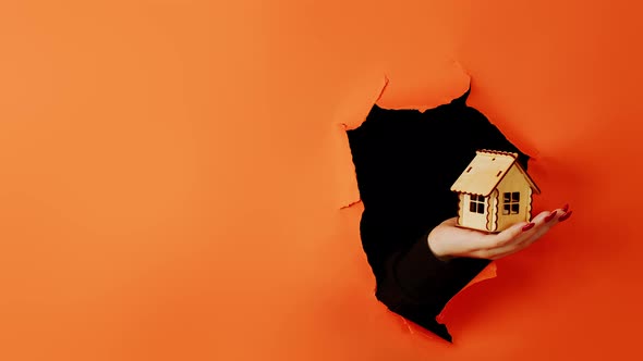 Close Up of Woman's Hand Holding Small Wooden House Through a Hole in the Orange Wall in the Studio