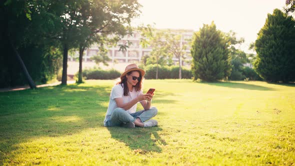 A Young Woman Uses Her Phone While Sitting on a Green Bright Lawn