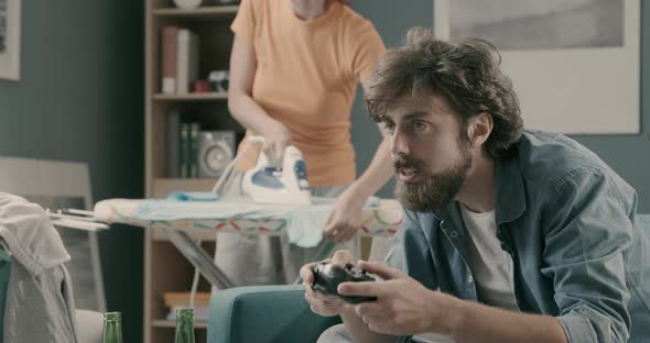 Lazy husband playing videogames while his wife is ironing