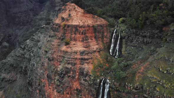 The drone rises above a rocky gray-red cliff and Waipoo Falls in Waimea Canyon, Kauai, Hawaii