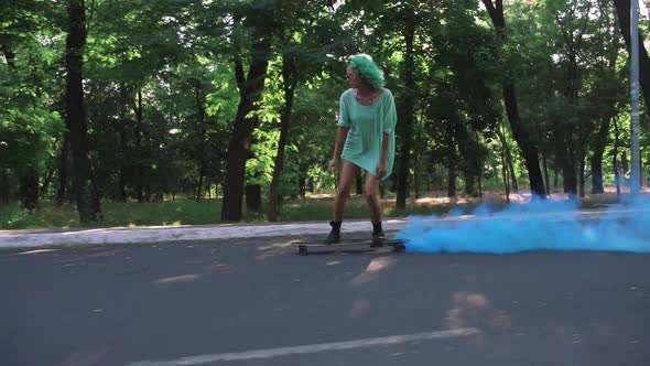 Beautiful Young Girl with Green Hair Riding on Longboard with Blue Smoke in Park Tracking Shot