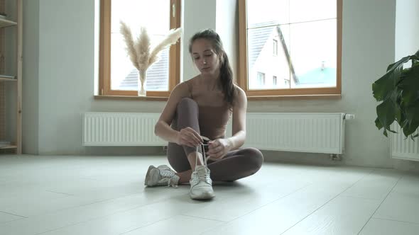 Young Girl in Sportswear Sitting on Floor on Knees Tying Shoelaces on Sneakers