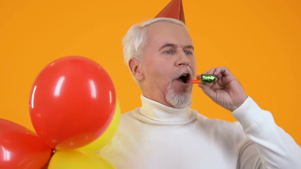 Joyful Senior Man Party Hat Blowing Noisemaker Holding Balloons, Celebration