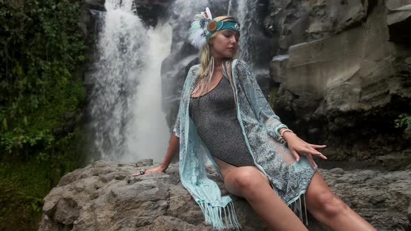 Enigmatic Young Lady Sitting on a Stone in Front of a Waterfall