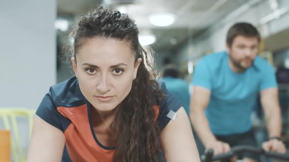 Close-up Portrait of Brunette Brown-eyed Woman Exercising on Exercise Bike with Blurred Man Riding