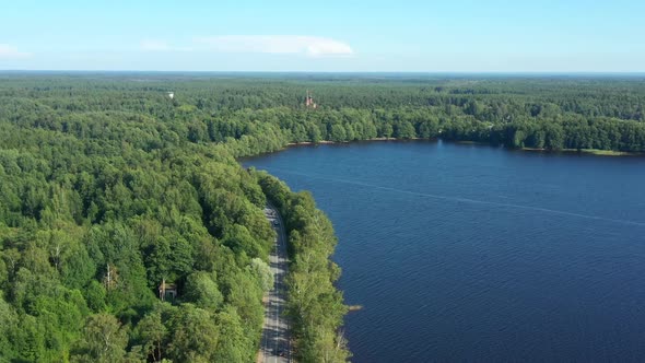 Asphalt Road with Driving Cars Through Green Summer Forest Near Lake. Aerial Shot From Drone