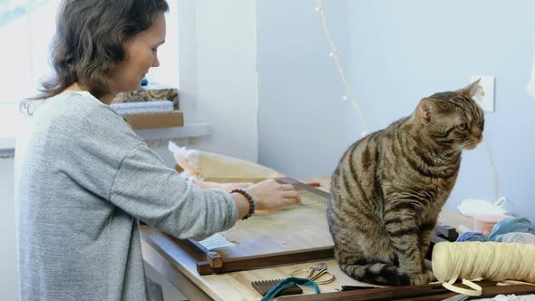 Woman Weaving on a Loom Frame. Cat Is Sitting Near Her on the Table.