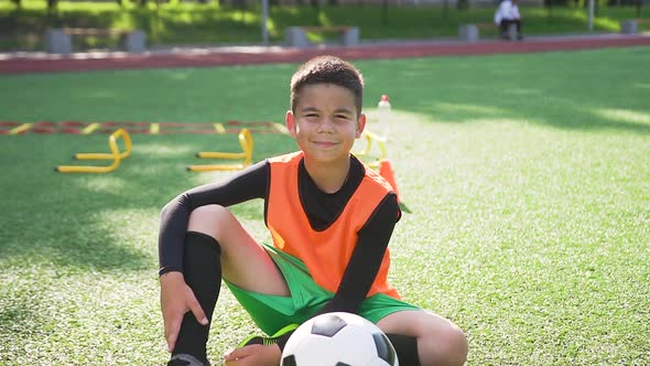 Teen Football Player in Uniform which Sitting with Ball on the Artificial Turf of Sport Field